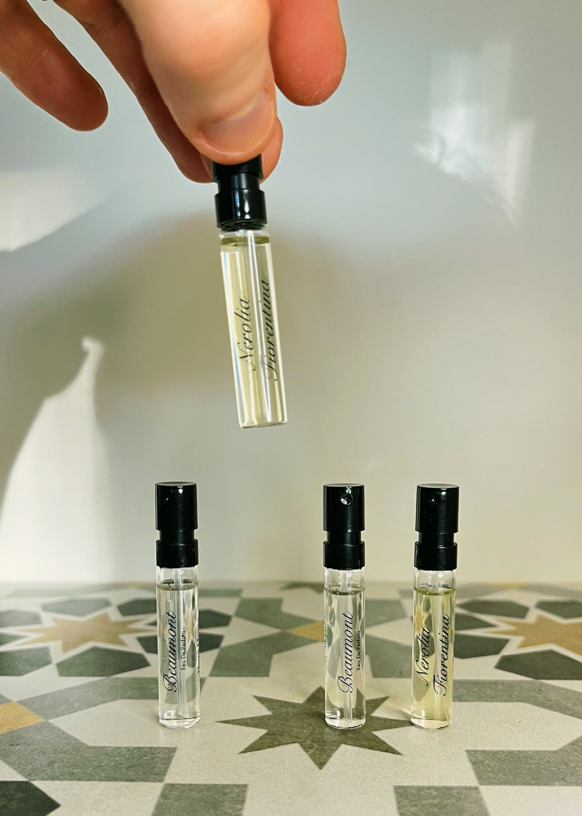 A hand holds a small clear perfume sample vial above three more vials standing on a patterned tile surface.